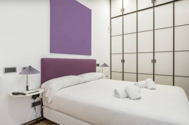 Bedroom with a double bed with a white duvet, pillows, a purple upholstered headboard and a custom-made wardrobe with white doors and wooden edges