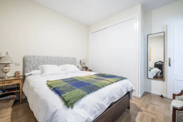 Bedroom with a double bed with an upholstered headboard and a white duvet with a blanket on it, a mirror hanging on the wall and a built-in wardrobe with white wooden sliding doors