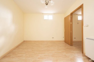 Empty living room with light yellow walls, light wooden floor, electric heater, oak wooden door, skylights high on the walls and ceiling lamp with white shade