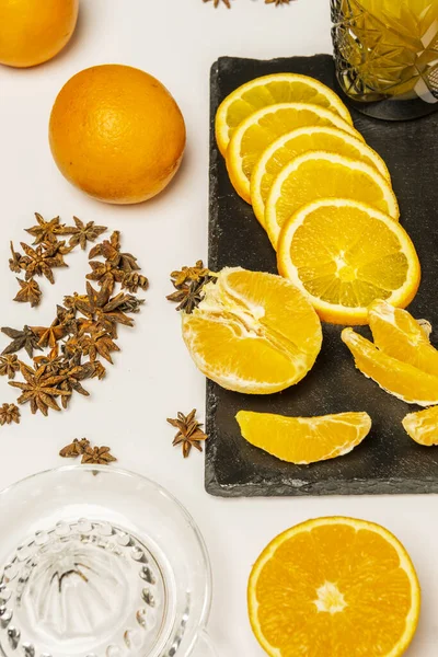 Some whole oranges with another peeled and another cut in half together with a quantity of star anise, a glass juicer and segments of fresh and succulent orange