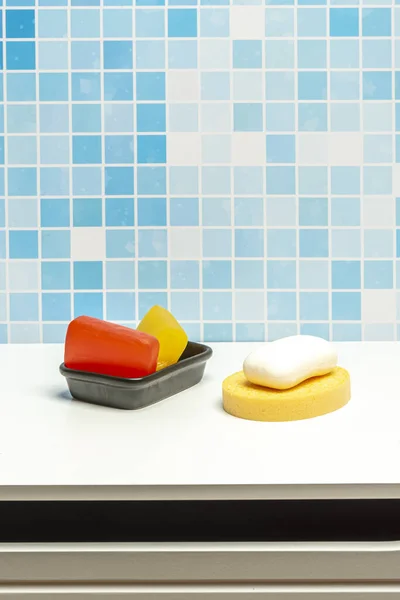 Black porcelain bowl with glycerin soap bars, a white soap bar and a yellow sponge on a white surface for hand washing in a blue tiled bathroom