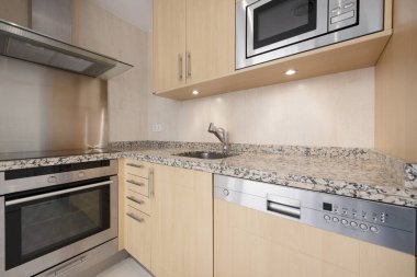 Corner of a newly installed modern kitchen with pink granite worktops, light wood cabinets, panel-integrated stainless steel appliances and courtesy lamps