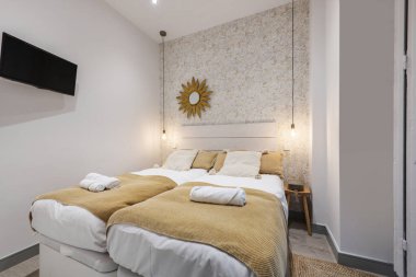 A bedroom with a double bed with white bedspreads and light mustard colored blankets with matching cushions and a wall with decorative wallpaper and a mirror with a gold metal frame