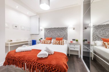 bedroom with a double bed with orange cushions and a gray wooden headboard, different bedside tables and a wardrobe with white sliding doors with a mirror