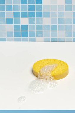 Yellow synthetic shower sponge on a white surface of a bathroom with foam and blue tiles on the wall