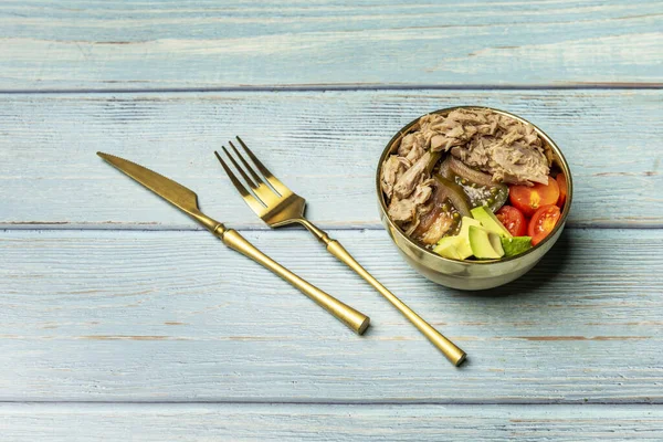 A salad with various kinds of diced tomato, avocado and lots of canned tuna in a golden bowl and matching cutlery