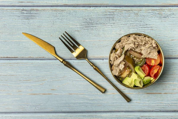 A wonderful canned tuna salad with diced tomatoes and avocados in a golden bowl with golden silverware to the side on a blue table