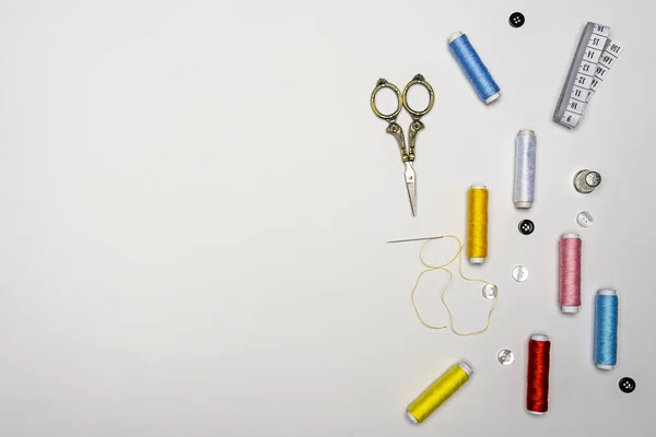 Set of spools of colored thread with a threaded needle, black and white buttons, golden scissors, white coiled tape measure on a white background