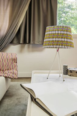 A space decorated with a patterned lamp on a white coffee table and in the background a sofa and a window with dark curtains