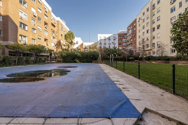 A summer pool covered with canvas to spend the winter in the common areas of an urbanization with green lawn gardens, trees, wooden benches and terrazzo floors