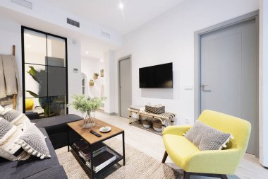 Living room with a three-seater sofa with a chaise longue in gray fabric and a glass partition on one wall, an industrial-style coffee table, mirrors leaning on the wall, a yellow armchair