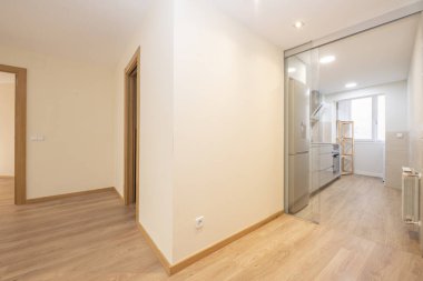 Corridor of an empty apartment and access to a kitchen furnished with smooth and shiny white cabinets with stainless steel handles, a wooden countertop and oak flooring