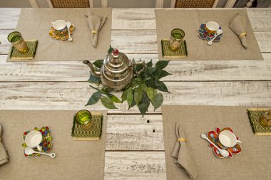 A white wooden dining table with small white porcelain teacups and metal teapots on a gray cloth tablecloth