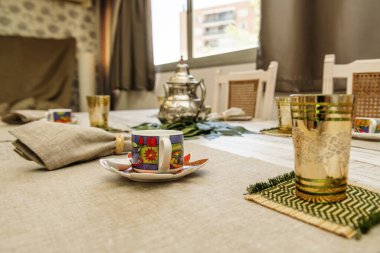 A small white porcelain cup with teacups and metal teapots on a gray cloth tablecloth
