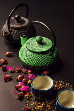 Still life with black and green Asian style cast iron teapots, small porcelain cups, green tea and dried rose tea