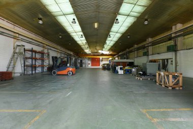 Metal raflarda ve kare kapılarda enerji ve enerji boruları olan sanayi deposu çinko çatılı çatılar, şeffaf ışıklar ve lambalar ve forklift.