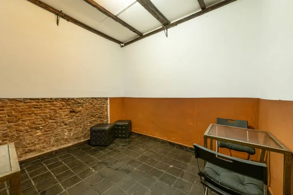 An almost empty room with black tile soils, brick wall and a metal table with two folding chairs and black square puffs