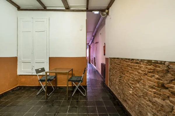 An almost empty room with black tile soils, brick wall and a metal table with two folding chairs