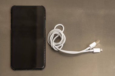 Gri metal bir masa ve beyaz şarj kablosu üzerinde karbon fiber kablosu olan siyah bir akıllı telefon.