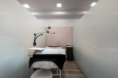 Sertleştirilmiş cam duvarlı estetik tedavi kabini masaj masası, çift katlı lambalar, siyah metal çekmece ve katlanmış döşeme.