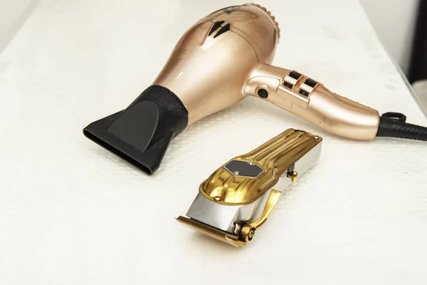 A golden hair dryer next to a hair clipper on a white surface