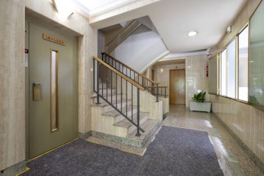 Zeminde halı, merdivenlerin yanında mermer fayanslar ve asansör olan bir iskan binasının iç kapısı.
