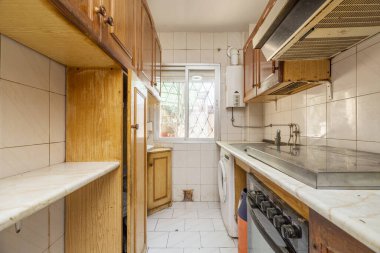 Çok kullanılmış ve yıpranmış ahşap dolapları ve arka planda alüminyum penceresi olan bir apartman mutfağı.