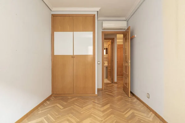 Empty room with a built-in wardrobe with two folding oak doors and a gloss varnished wooden floor placed in a herringbone pattern and a matching access door