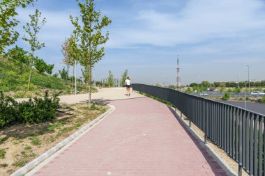 Yeni bir şehir parkında kırmızı tuğla kaldırım ve metal korkuluklarla koşu yapan biri.
