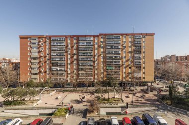 Güneşli bir günde parkları ve otoparkları olan büyük açık alandaki binaların ana cepheleri.