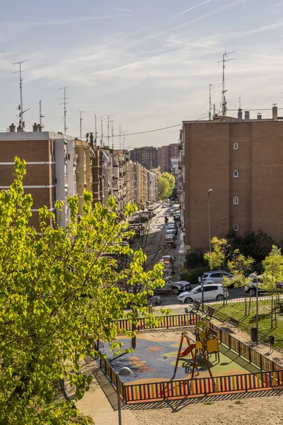 Şehir caddesindeki binaların manzarası ve ön planda bir oyun alanı, bahçeler ve ağaçlar.