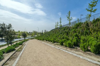 Çiçek açan bitkiler, çitler ve genç ağaçlarla çevrili kumlu bir şehir parkı.