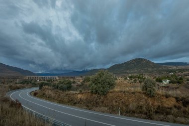 Alçak çalılı Sierra de Madrid manzarası, fırtına bulutlarının olduğu bir günde dolambaçlı bir yol.