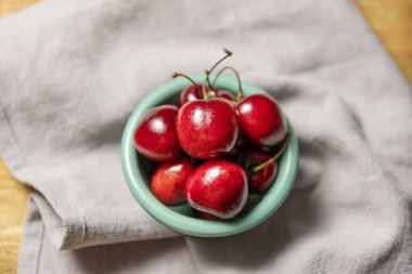 Küçük yuvarlak yeşil porselen kâse, üzerinde olgun kirazlar, gri kumaş sapları ve verniklenmemiş ahşap arka planı var.