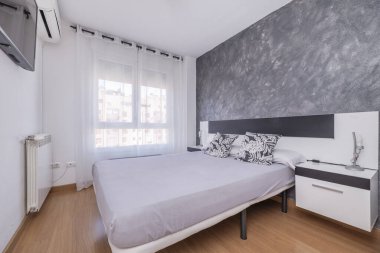 Kantilever komodinli yorganı olmayan çift kişilik bir yatak odası, siyah beyaz yatak başlığı, yarı şeffaf beyaz perdeli bir pencere ve alüminyum bir radyatör.