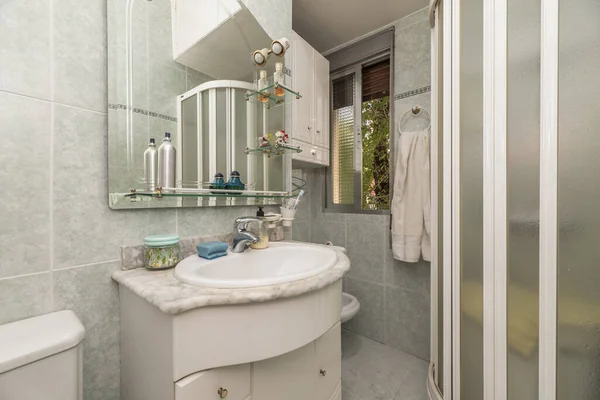 Small bathroom with porcelain sink on marble countertop with small white cabinet with mirror and a shower cabin