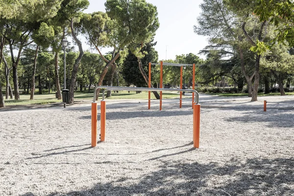 Orange gym equipment in a park with a gravel surface and lots of leafy trees
