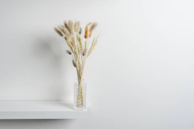 Dikenli küçük cam vazo ve kurumuş bitki dalları, bazıları beyaz ahşap bir rafta ve düz beyaz duvarda boyalı.
