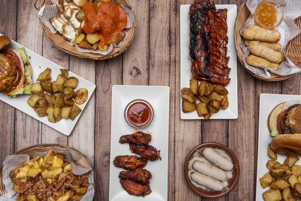 Set of international fast food dishes, barbecue ribs, burgers, patatas bravas, chicken wings with sauce and french fries with bacon and cheese, all very healthy on top of a wooden table