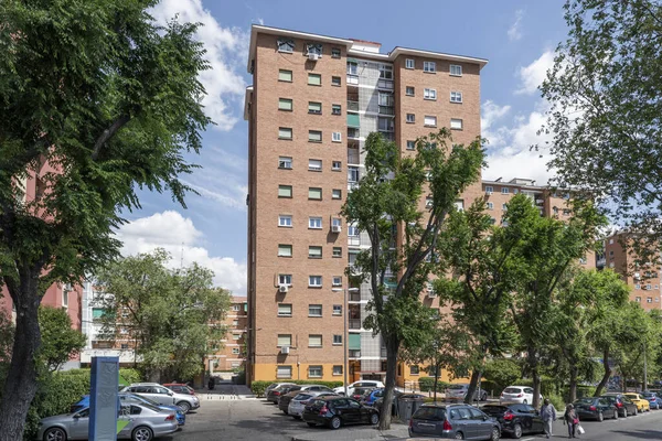 Mavi yay gökyüzünde bulutlar ve caddelerde yapraklı ağaçlar olan apartman cephesi.
