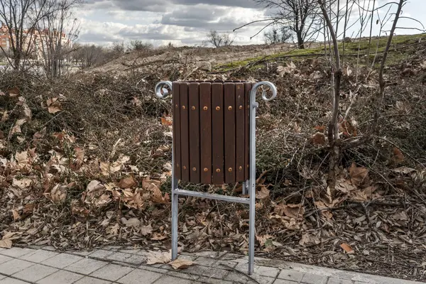 A metal and wooden trash can in a public park on a winter day with many fallen leaves