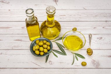 Birkaç şişe dolusu İspanyol zeytinyağı ve yeşil bir tabakta biraz zeytin, zeytin yaprakları ve beyaz ahşap bir masada yağlı bir kap.