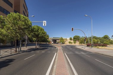 İki yönlü bir cadde. Ağaçlarla çevrili bir kavşak ve trafik ışıkları var.