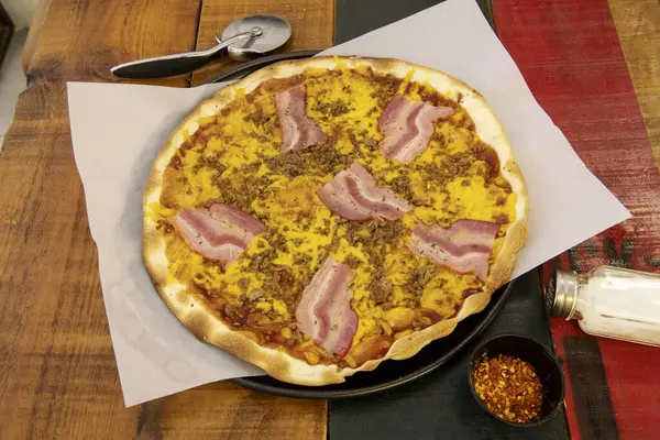 A wonderful medium thin crust pizza with bacon, various meats, cheddar cheese and pomodoro on a colorful wooden table