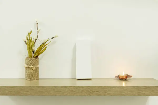 A light oak imitation wood shelf with a white cardboard package, an ornament of dried flowers and a small lit candle