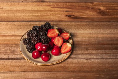 Olgun böğürtlen, çilek ve çiğ ahşap arka planda sapsız kiraz içeren küçük bir meyve tabağı.