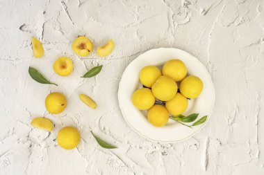 Beyaz bir porselen tabakta sarı erik ve pürüzlü beyaz bir yüzey üzerinde yeşil yapraklar.