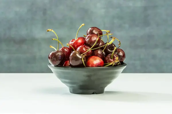 A small bunch of ripe cherries with stems inside a black bowl on a smooth blue surface
