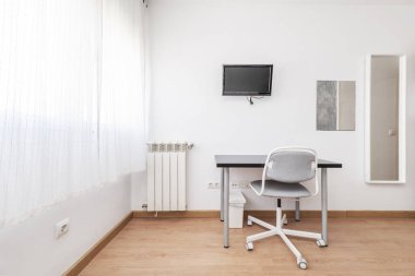 Bir duvarında beyaz perdeler ve döner sandalyeli küçük gri bir masa olan bir oda.