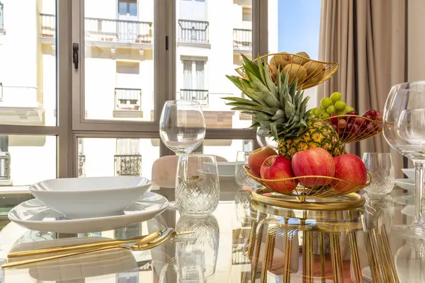 A glass dining table with a gold fruit bowl with pineapple, various types of grapes, kiwi, red apples, gold cutlery and crystal goblets by a window with a view